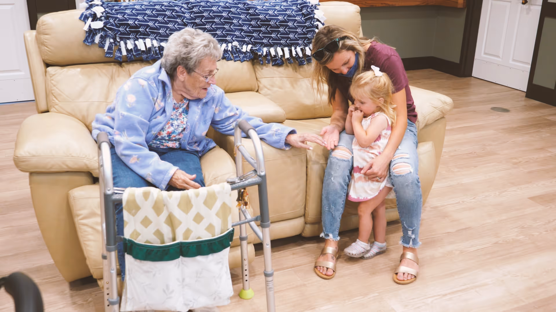 An elderly woman sitting on a beige leather couch reaching out her hand to a young woman and a little girl who are sitting and standing next to the couch in a cozy indoor setting with wooden flooring. A walker with a hanging organizer is in front of the elderly woman.