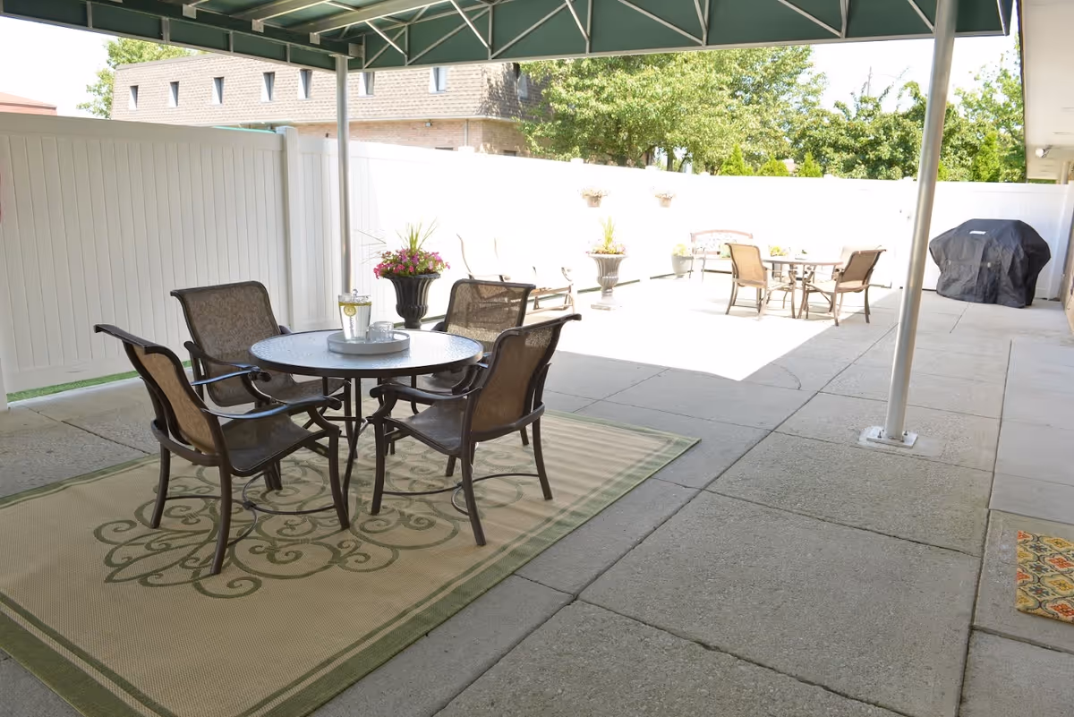 Outdoor patio area with two sets of metal tables and chairs on concrete flooring. One table is under a green canopy with a decorative rug underneath, and the other is in the open sun. There are potted plants and a covered grill along the white fence surrounding the patio.