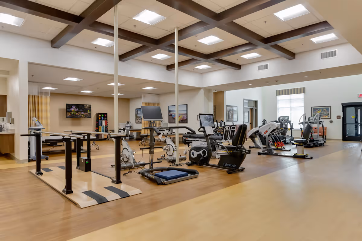A spacious fitness room in a senior living facility with various exercise equipment including stationary bikes, a treadmill, parallel bars for walking support, and other workout machines. The room has a high ceiling with wooden beams and bright overhead lighting. There are windows and framed pictures on the walls, and the floor is a combination of light and medium-toned wood.