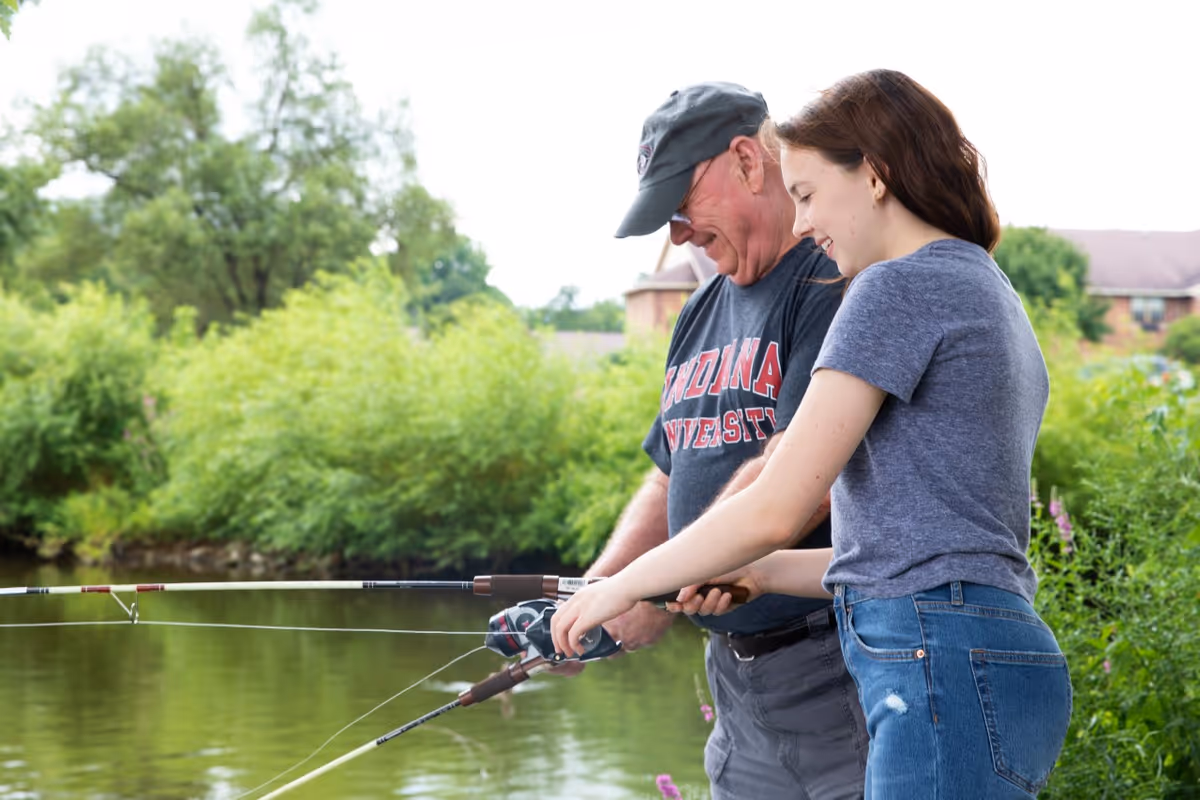 An older man and a young woman fishing together by a pond surrounded by green bushes and trees. The man is wearing a dark cap and an Indiana University t-shirt, while the woman is smiling and helping with the fishing rod.