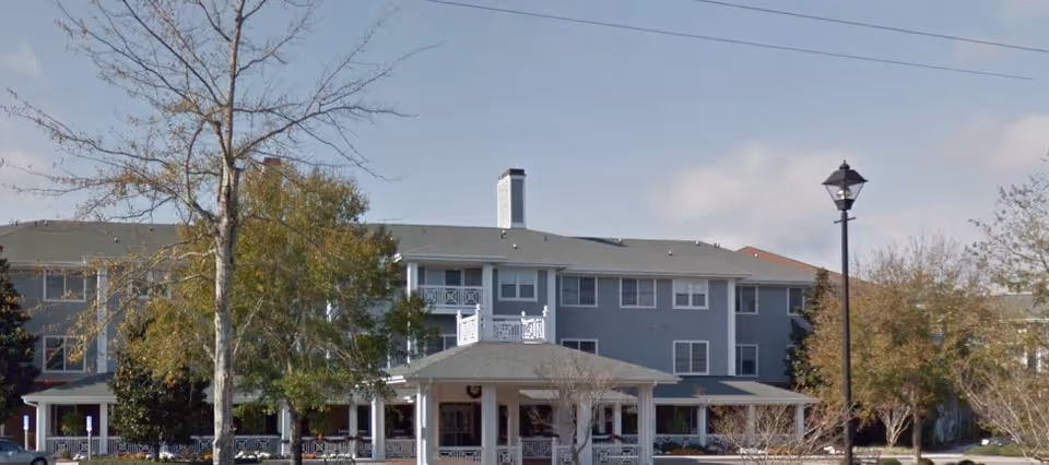 Exterior view of a multi-story senior living facility building with a covered entrance, surrounded by trees and a street lamp under a partly cloudy sky.
