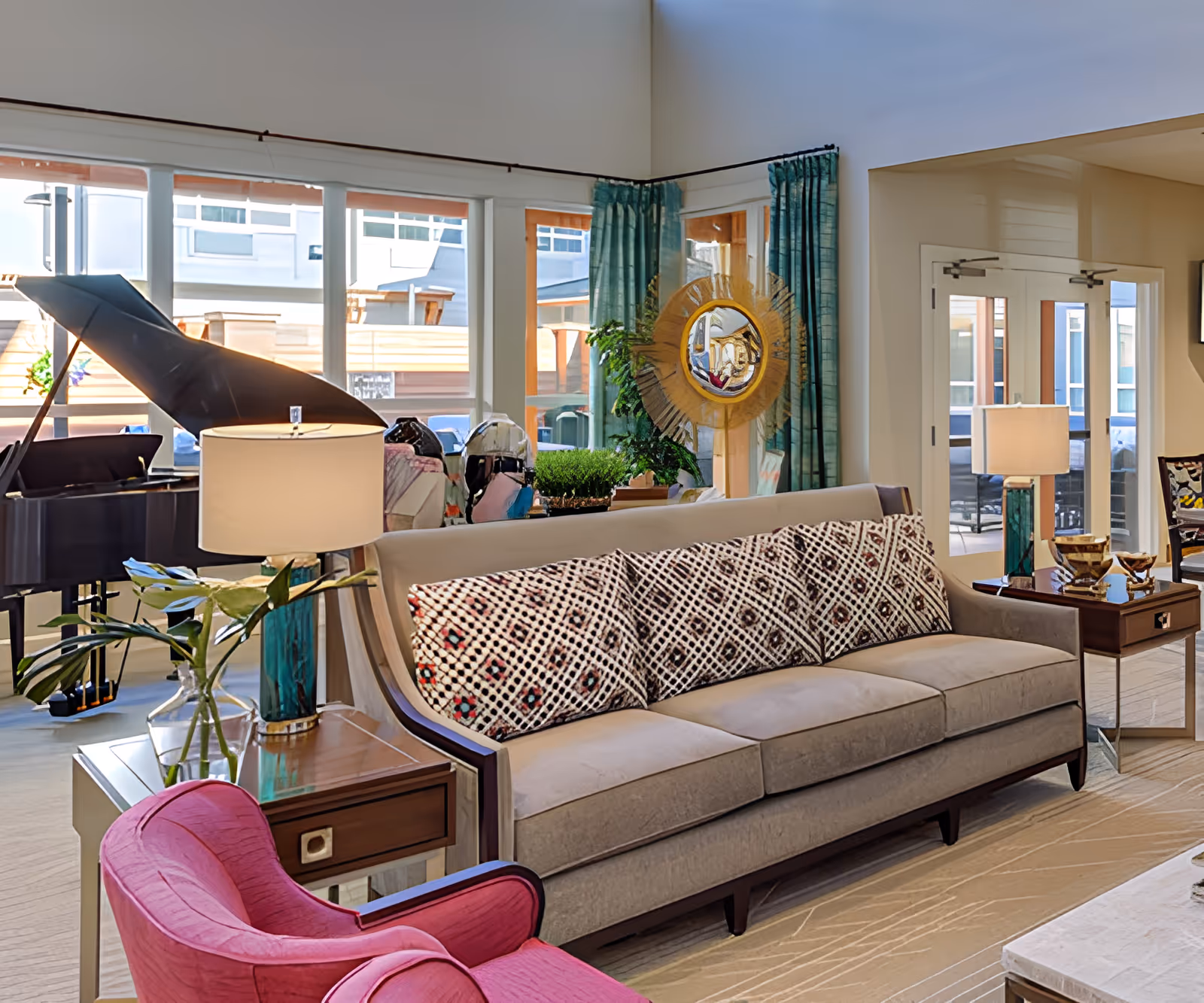 Bright assisted living common room with a sofa and patterned pillows, pink armchair, grand piano, lamps, and large windows.