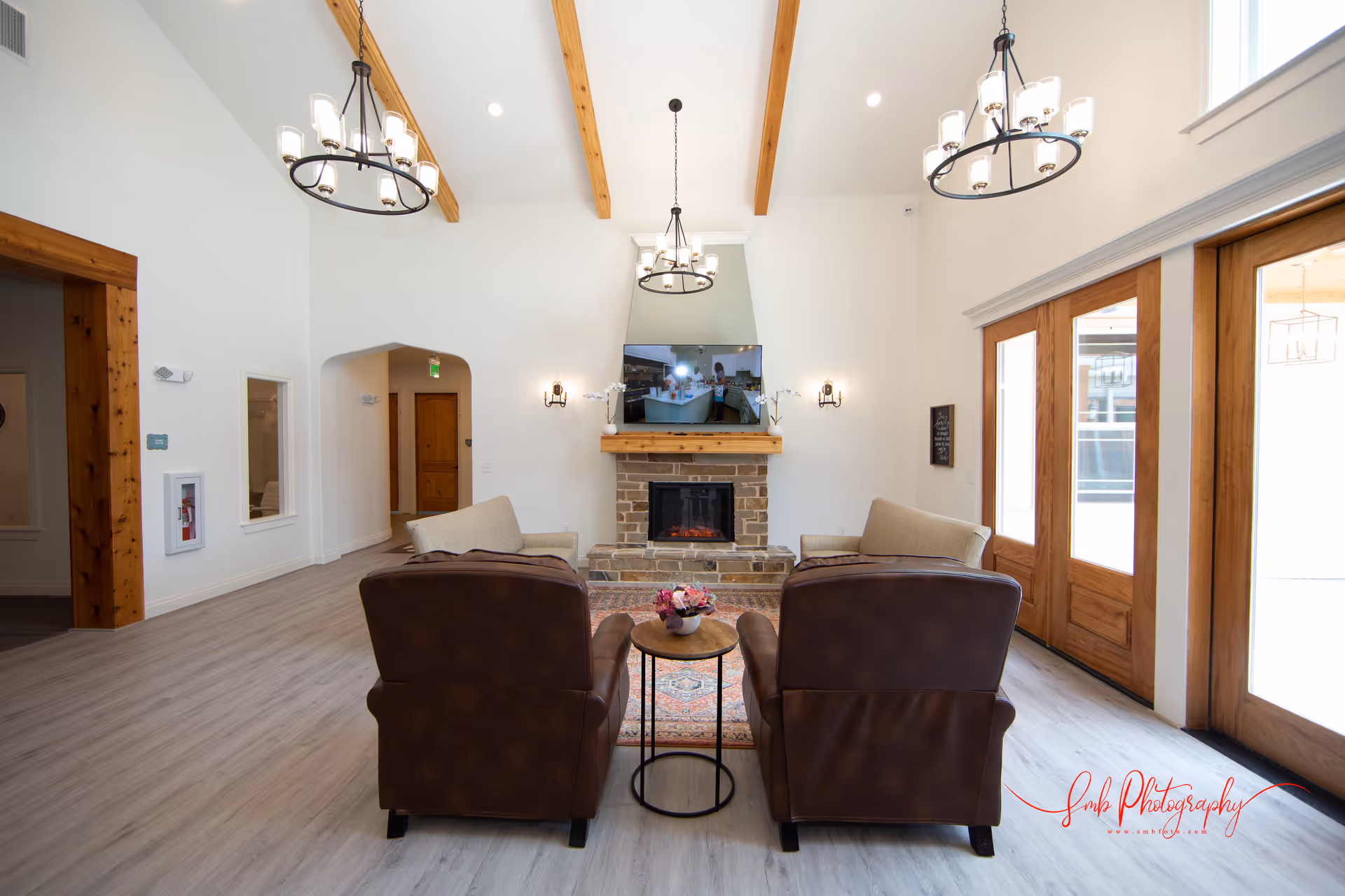 A bright and spacious living room with high ceilings and wooden beams. The room features two brown leather armchairs and two beige sofas arranged around a small round wooden table with a flower vase. A stone fireplace with a mounted flat-screen TV is centered on the far wall. Large wooden-framed glass doors allow natural light to fill the space. Modern chandeliers hang from the ceiling, and light wood flooring complements the neutral color scheme.