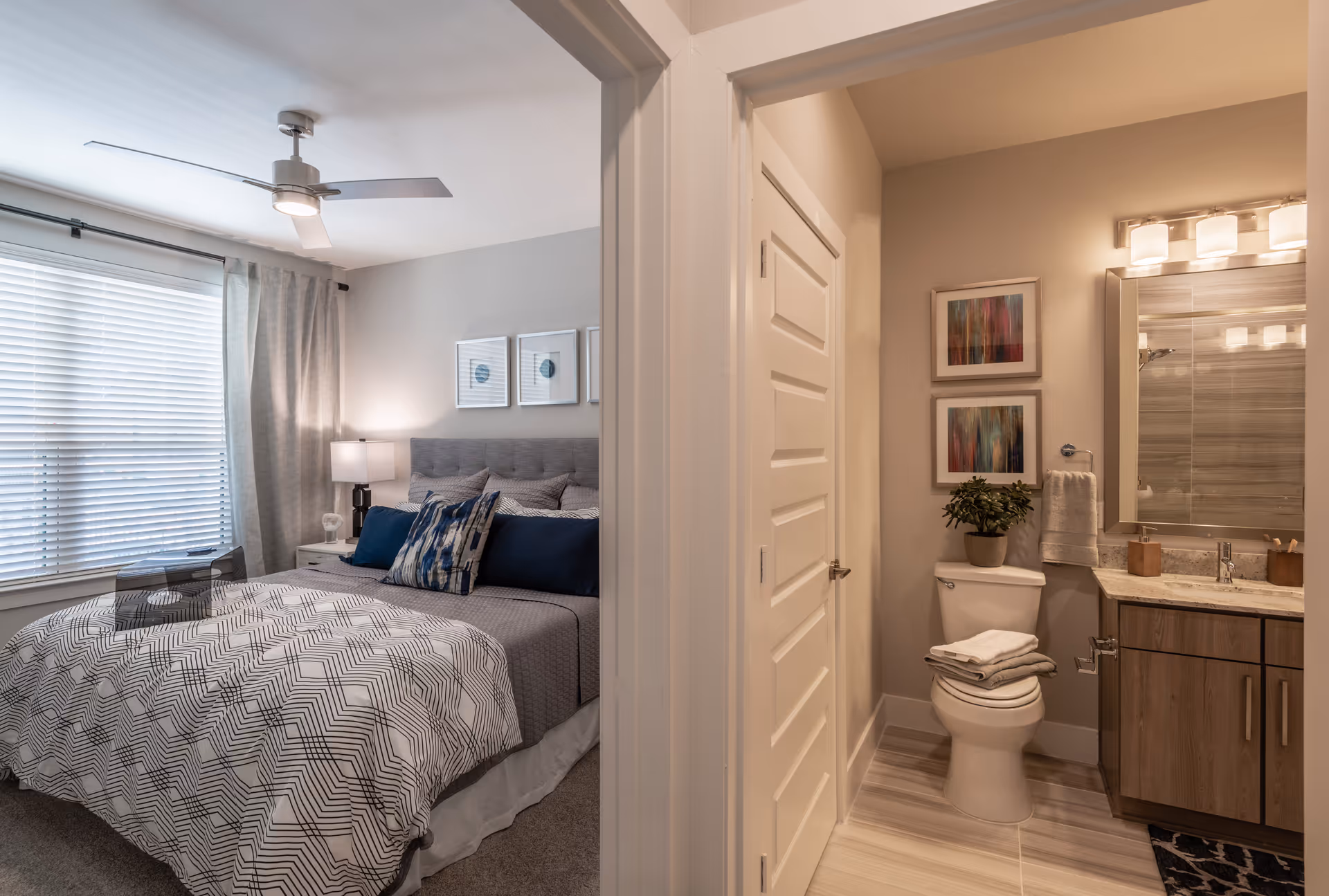 A cozy bedroom with a neatly made bed featuring patterned bedding and multiple pillows, next to a window with blinds and curtains. Adjacent to the bedroom is a bathroom with a toilet, a vanity with a sink, a mirror with lights above it, and two framed artworks on the wall.