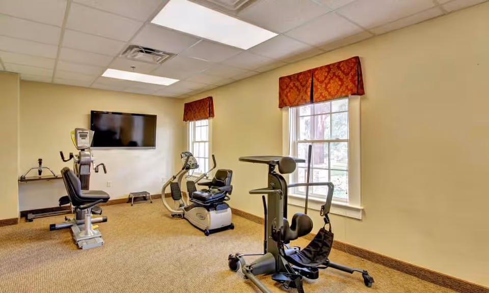 A small exercise room with beige walls and carpeted floor, containing three pieces of exercise equipment including a recumbent bike and a seated resistance machine. Two windows with red patterned valances allow natural light into the room, and a flat-screen TV is mounted on the wall.
