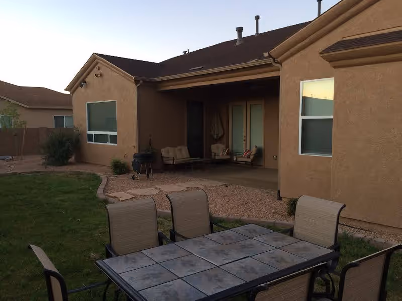 Backyard patio area of a residential building with a tiled outdoor dining table and six chairs on a grassy lawn. The patio has a covered seating area with cushioned chairs and a small table, along with a barbecue grill. The building exterior is tan stucco with several windows and a set of French doors leading to the patio.