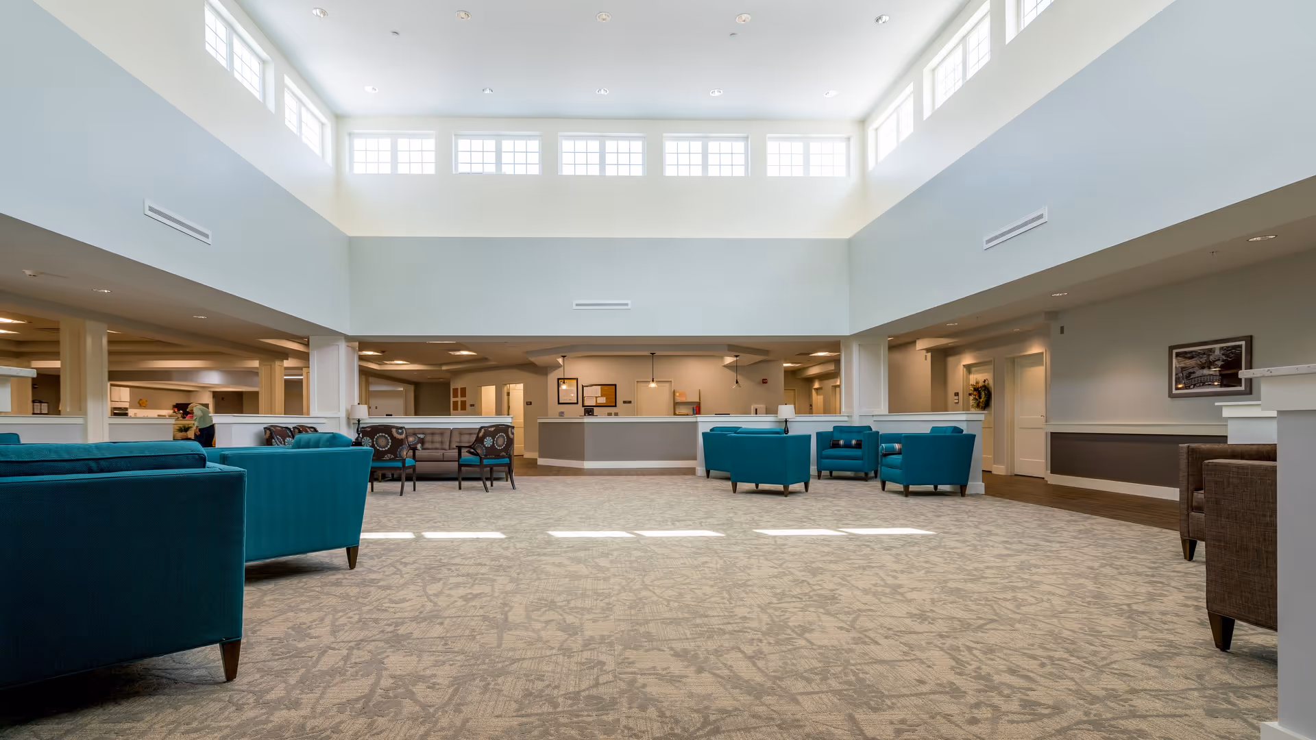 Spacious and well-lit common area in a senior living facility with high ceilings and large windows near the ceiling allowing natural light to fill the room. The room features multiple seating arrangements with teal and brown upholstered chairs and sofas placed on a patterned carpet. The walls are painted light gray with white trim, and there is a reception desk or service counter in the background. The overall atmosphere is calm and welcoming.
