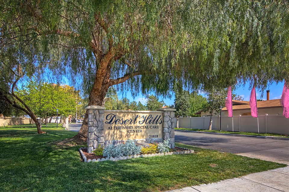 Stone sign for Desert Hills Alzheimer's Special Care Center surrounded by green grass and plants, with a large tree overhead and a paved driveway to the right under a clear blue sky.