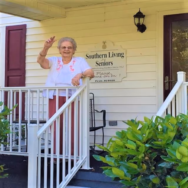 An elderly woman standing on the porch of a building, waving and smiling. Behind her is a sign that reads 'Southern Living For Seniors' with an invitation to come on in. The porch has white railings and there are green bushes in the foreground.