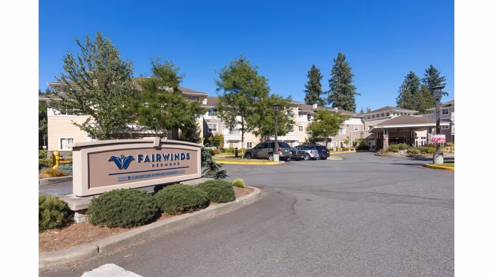 Exterior view of Fairwinds Redmond senior living facility with a large sign in the foreground, several trees, parked cars, and a multi-story building under a clear blue sky.