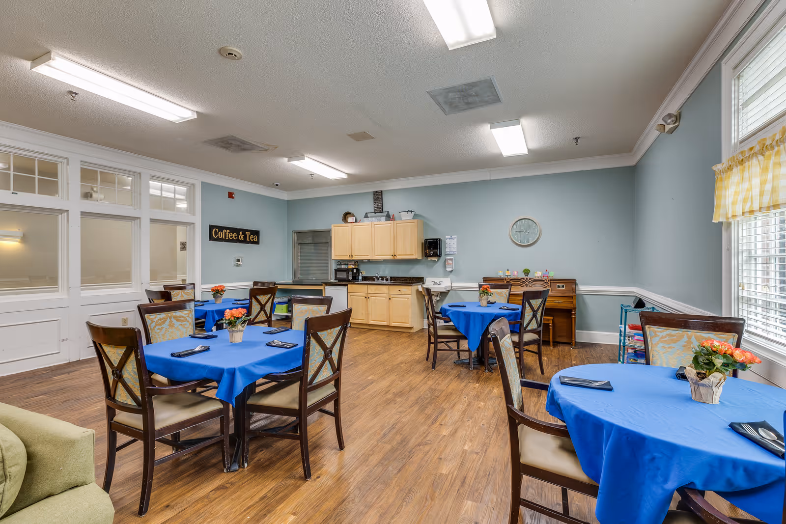 A dining area in an assisted living facility with several round tables covered in blue tablecloths, each set with black napkins and small flower arrangements. The room has wooden flooring, light blue walls, and a kitchenette with light wood cabinets in the background. A sign on the wall reads 'Coffee & Tea'. There is a window with yellow checkered curtains letting in natural light.