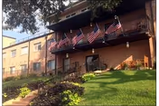 Exterior view of a multi-story brick building with a balcony displaying several American flags. There is a grassy slope with a garden and a ramp leading to the entrance.