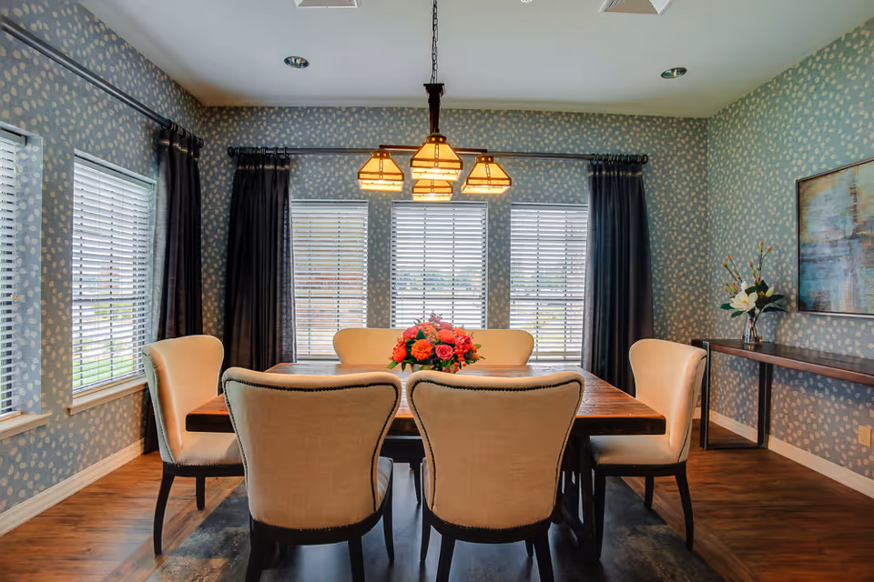 A dining room with a wooden table surrounded by six beige upholstered chairs. A floral centerpiece is on the table. The walls are decorated with blue wallpaper featuring a subtle white pattern. Three large windows with white blinds and dark curtains allow natural light into the room. A modern chandelier with four lights hangs above the table. A side table with a vase of flowers and a framed abstract painting are visible on the right wall.