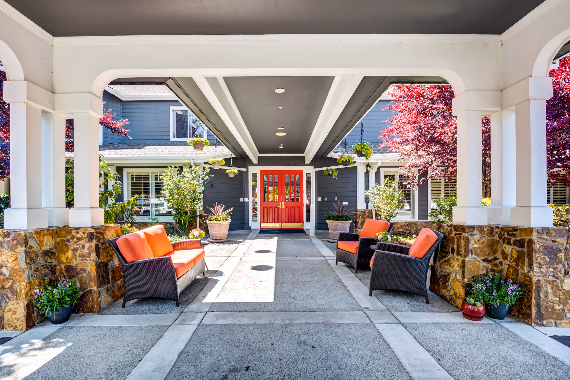 Covered outdoor seating area with stone pillars and white beams, featuring wicker chairs and a loveseat with orange cushions. The area leads to a building entrance with double red doors, surrounded by plants and trees with red foliage.