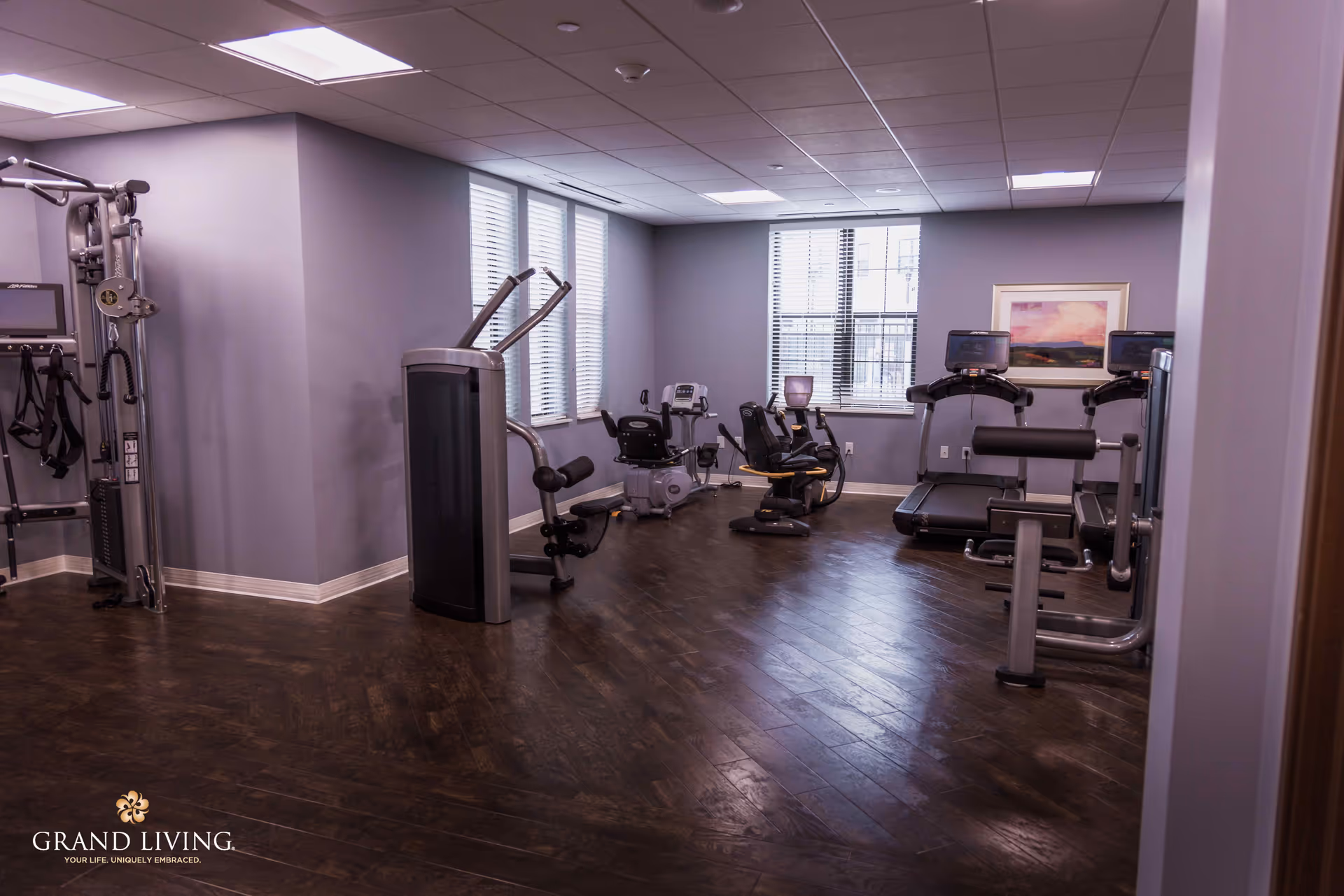 A fitness room with various exercise equipment including treadmills, stationary bikes, and weight machines. The room has wooden flooring, gray walls, and windows with blinds allowing natural light to enter. A framed picture hangs on the wall.