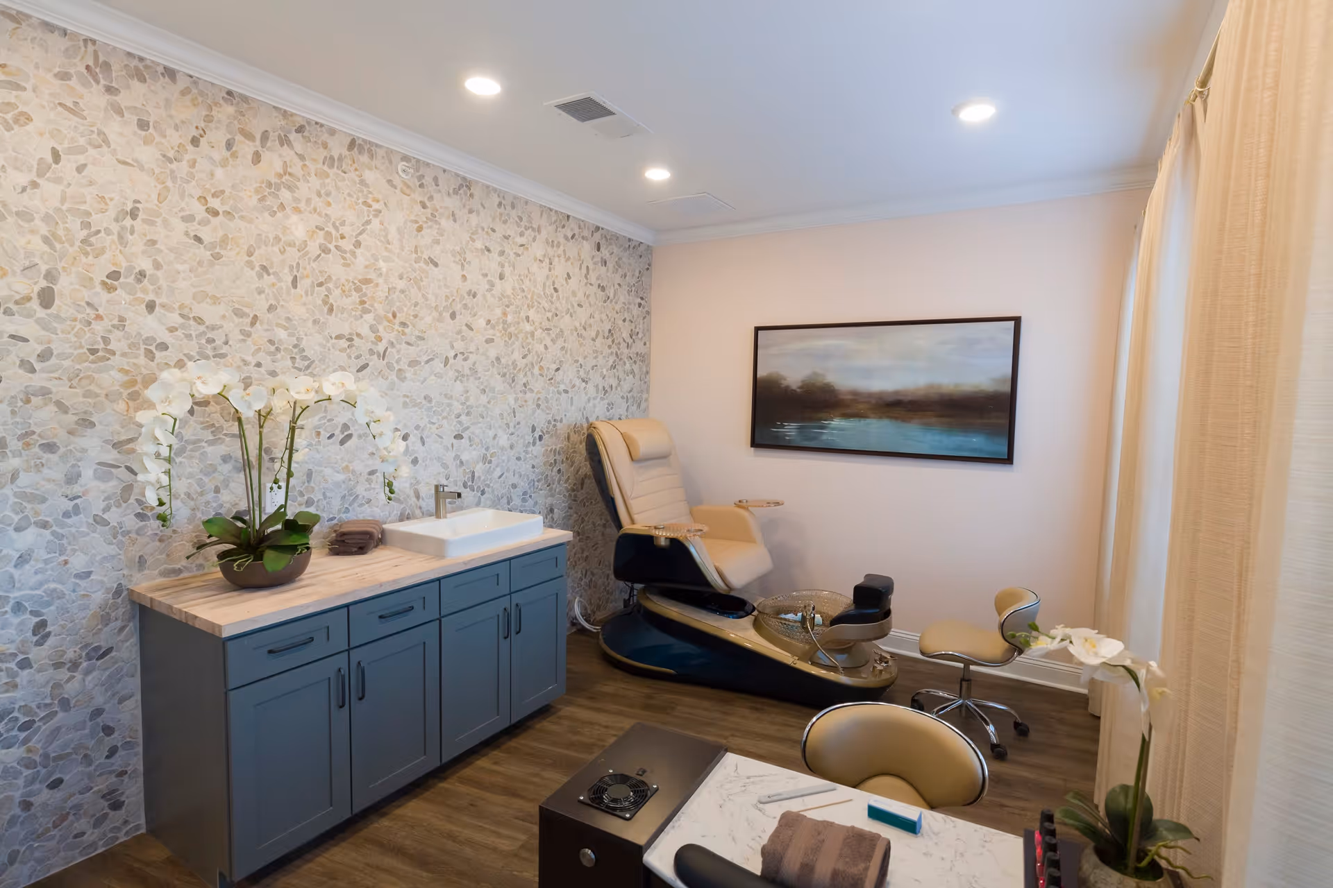 A well-lit room with a pedicure spa chair, a small rolling stool, a cabinet with a sink and a potted orchid on top, and a framed landscape painting on the wall. The room has a pebble-textured accent wall, wood flooring, and light-colored curtains covering a window.