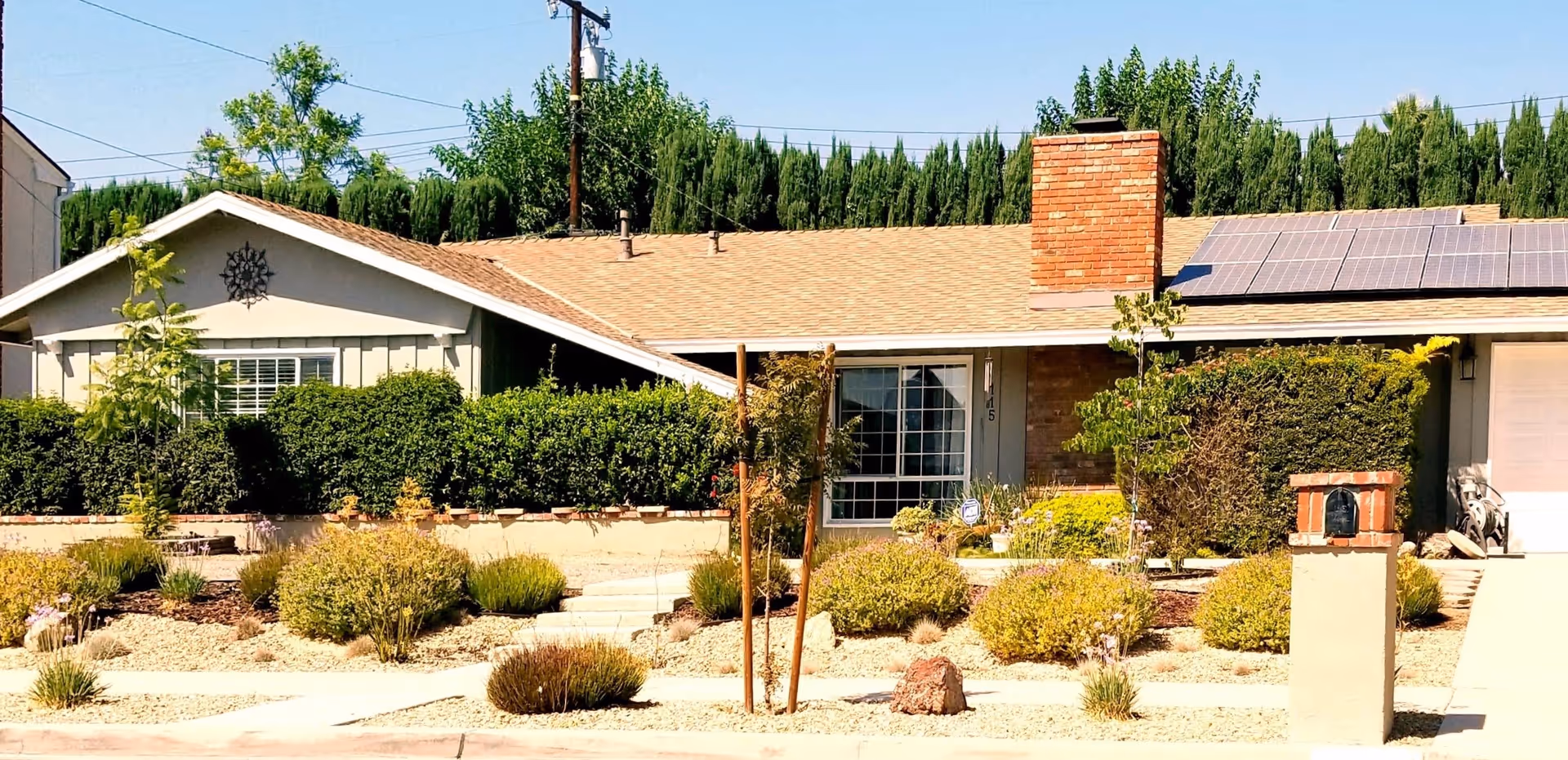 Single-story residential building with a beige roof, solar panels, a brick chimney, and a well-maintained front yard featuring various shrubs, small trees, and a stone pathway leading to the entrance.