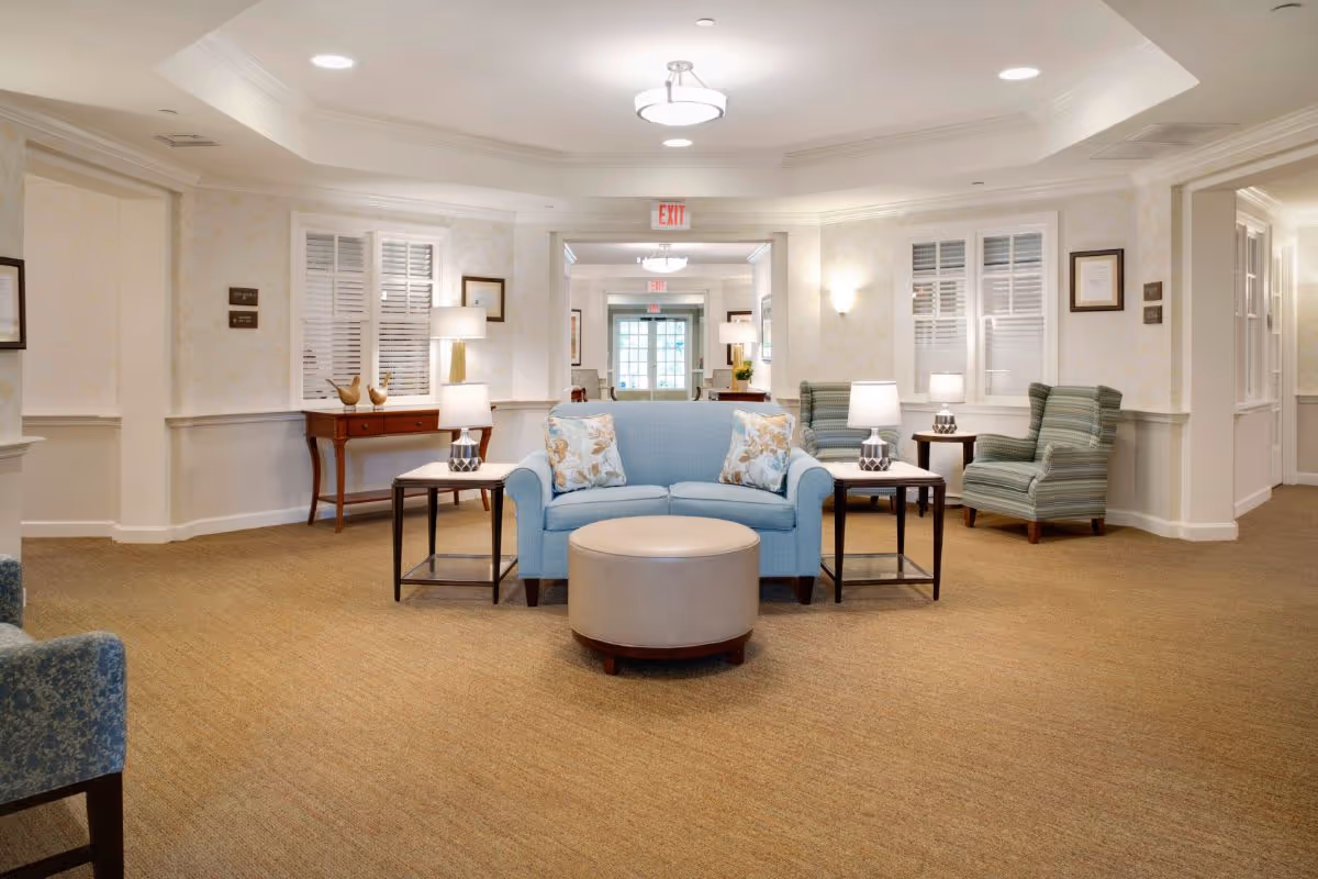A well-lit senior living facility common area with a light blue loveseat, two striped armchairs, a round beige ottoman, and side tables with lamps. The walls are light-colored with white trim and windows with white shutters. There is a wooden console table with decorative items and framed pictures on the walls. The carpet is beige, and there are exit signs visible in the background.