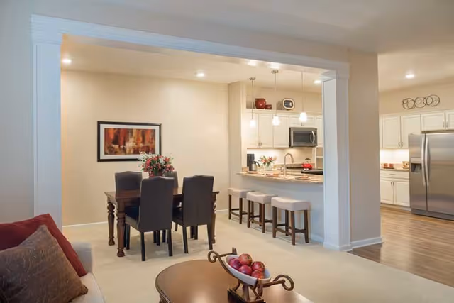 Open-concept living area with a coffee table in the foreground, a dining table and chairs, and a kitchen island with stools and stainless steel appliances in the background.