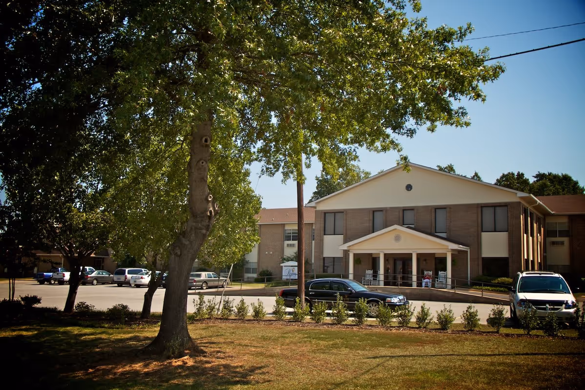 Exterior view of a two-story brick building with a covered entrance supported by columns. There are rocking chairs on the porch and several cars parked in front. A large tree with green leaves is in the foreground, and the sky is clear and blue.