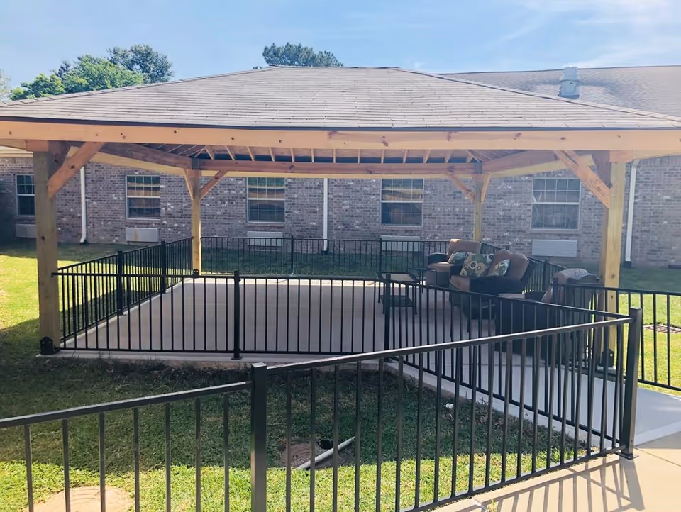 Outdoor covered seating area with a wooden roof structure and black metal railing. The area contains cushioned chairs and a small table, situated on a concrete platform with grass surrounding it. In the background, there is a brick building with multiple windows and air conditioning units.