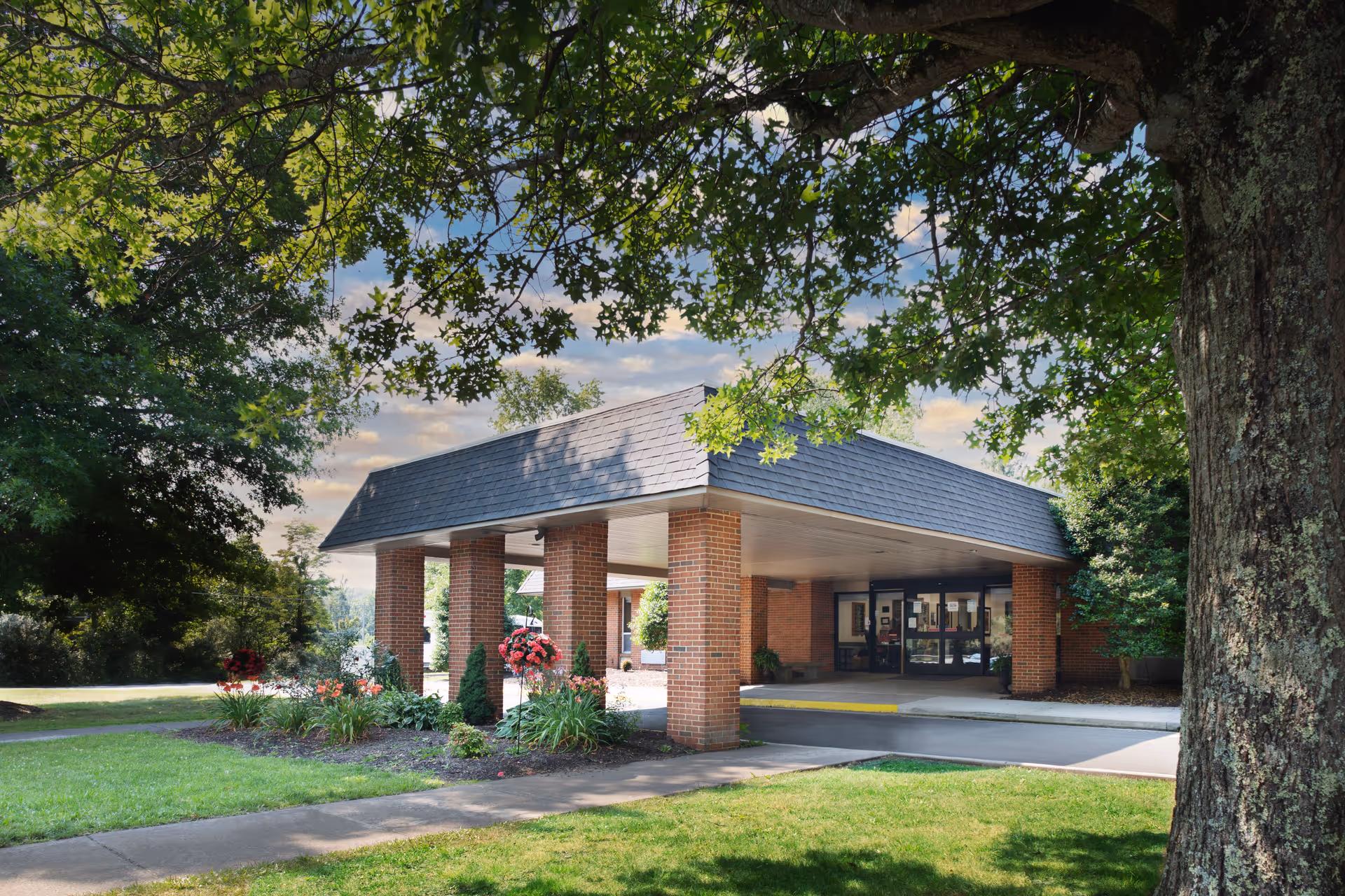Entrance of a brick building with a covered driveway supported by brick columns, surrounded by green grass, trees, and flower beds under a partly cloudy sky.