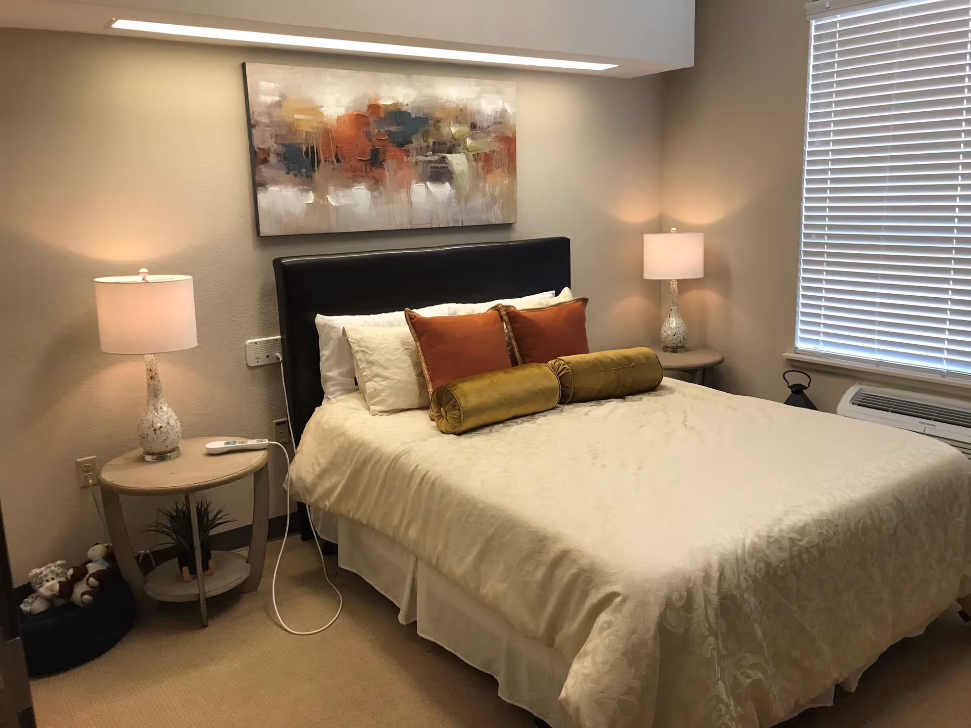 Bright furnished bedroom with a made bed, decorative pillows, two bedside tables with lamps, wall art above the headboard, and a window with blinds.