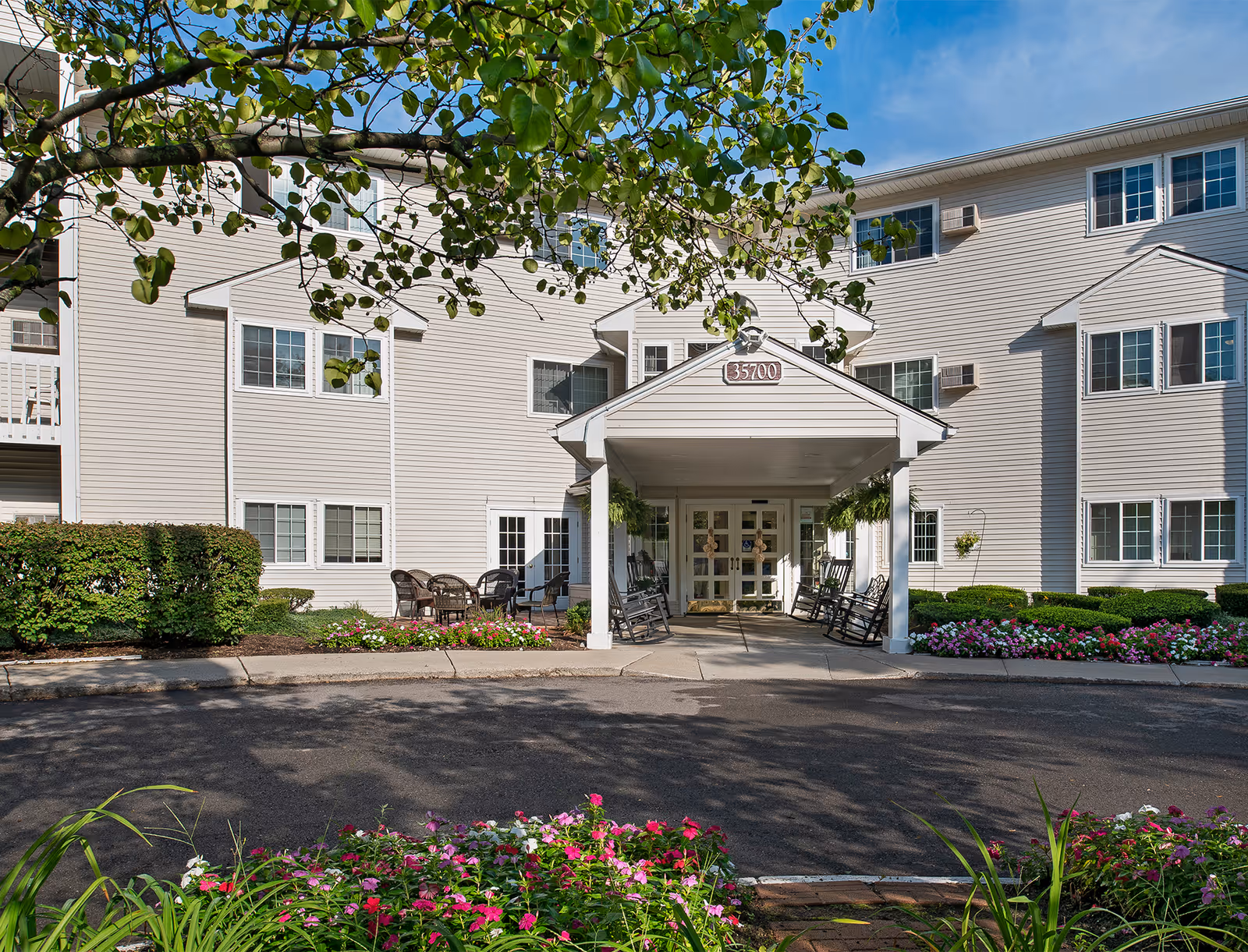 Front entrance of a senior living facility with a covered porch, rocking chairs, outdoor seating, and well-maintained flower beds and shrubs under a clear blue sky.