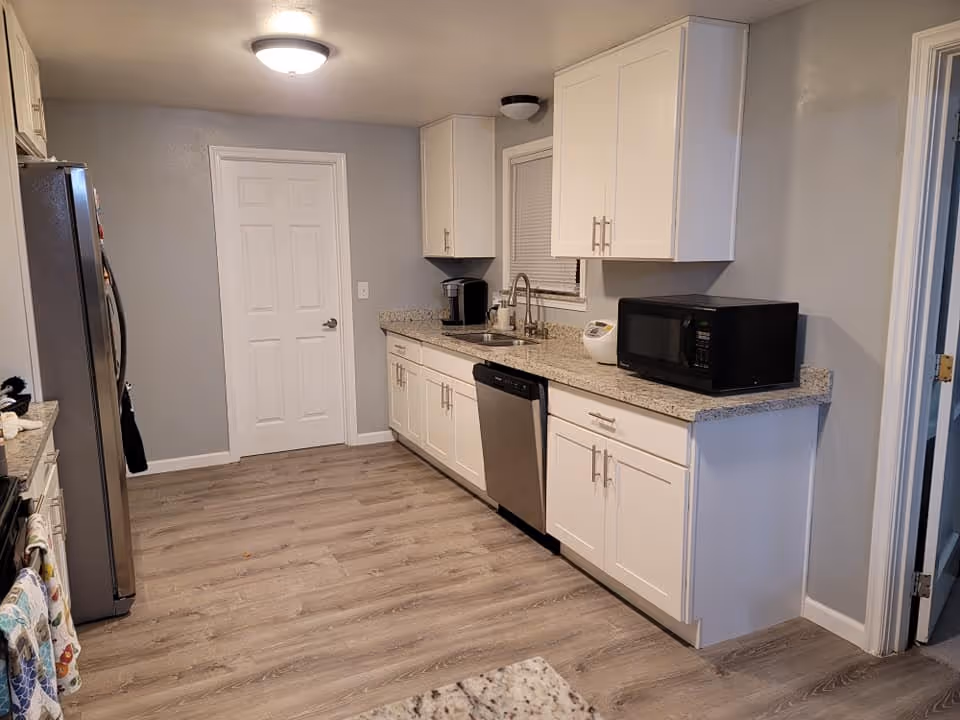 Modern galley kitchen with white cabinets, granite countertops, a stainless steel dishwasher and refrigerator, microwave on the counter, and wood-look flooring.