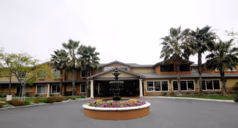 Front exterior view of Cypress Place facility with a circular driveway featuring a central fountain surrounded by flowers, palm trees, and a two-story building under an overcast sky.