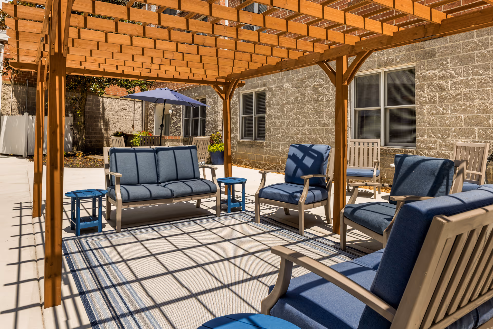 Outdoor patio area with a wooden pergola casting shadows on a seating arrangement consisting of cushioned chairs and a sofa in blue fabric, small blue side tables, and a dining table with chairs and a blue umbrella in the background, adjacent to a stone building with windows.