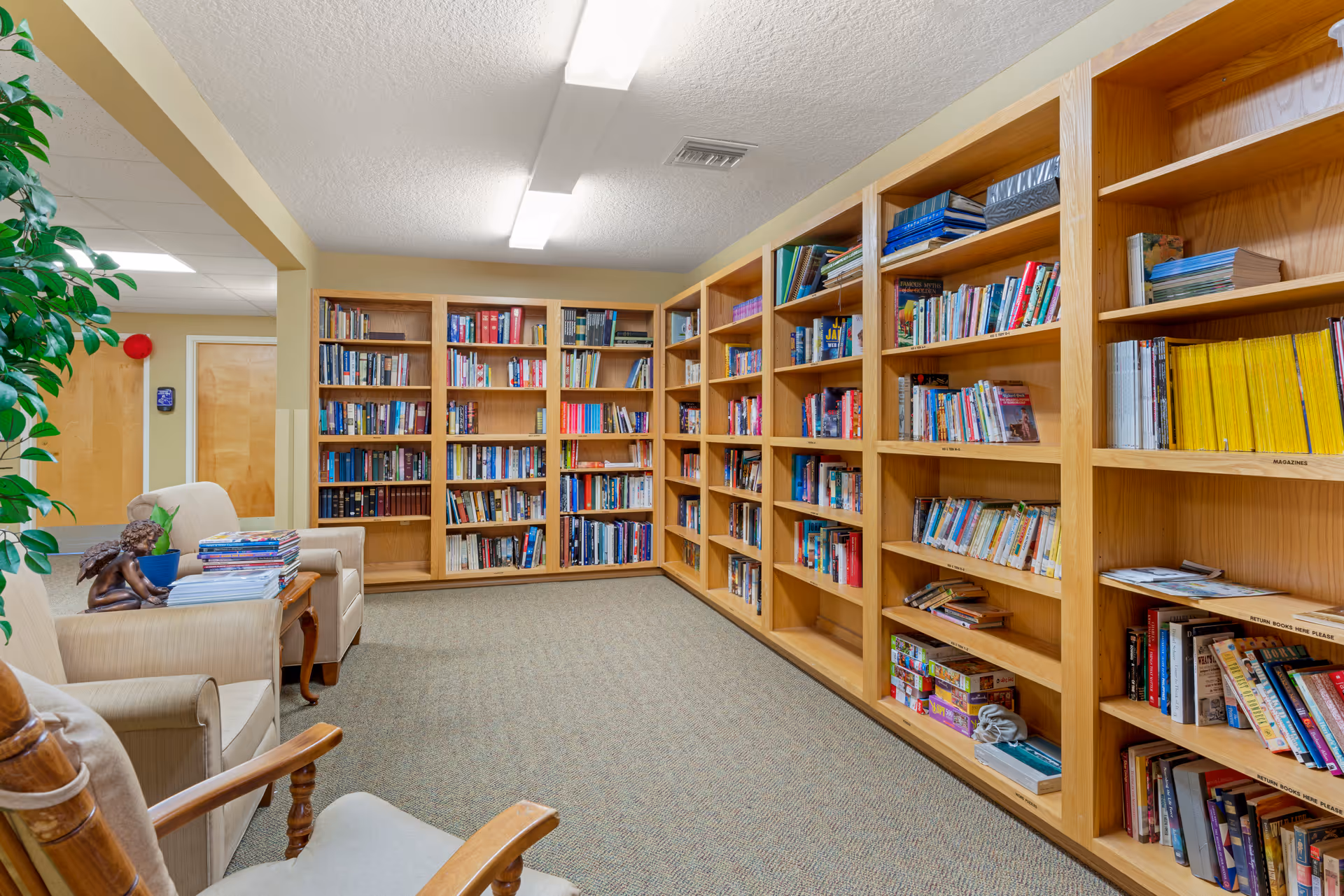 A cozy library room with wooden bookshelves filled with books and magazines lining two walls. There are comfortable armchairs and a small table with a decorative sculpture and a stack of magazines. The room has carpeted flooring and bright overhead lighting.