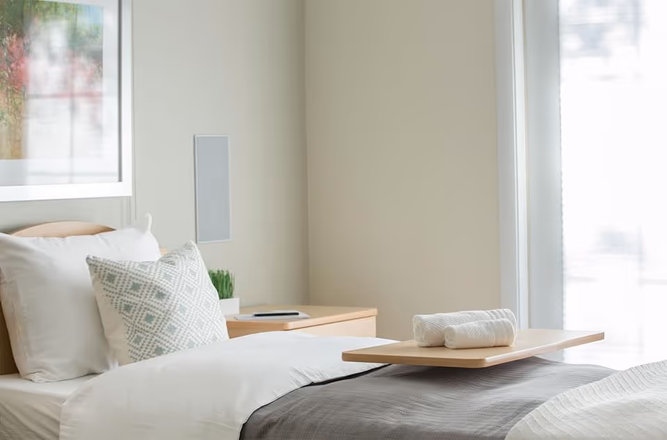 A bright, minimalist bedroom with a made bed, patterned pillow, bedside table and a tray holding folded towels.