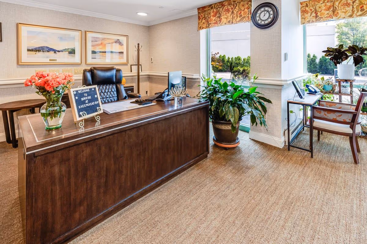 Reception area with a wooden desk, a black leather office chair, a vase of pink flowers, a welcome sign that reads 'Welcome to Sunrise at Sheepshead Bay', two framed landscape paintings on the wall, a coat rack, large green plants near windows with floral valances, a wall clock, and a small desk with a chair and phone by the window.
