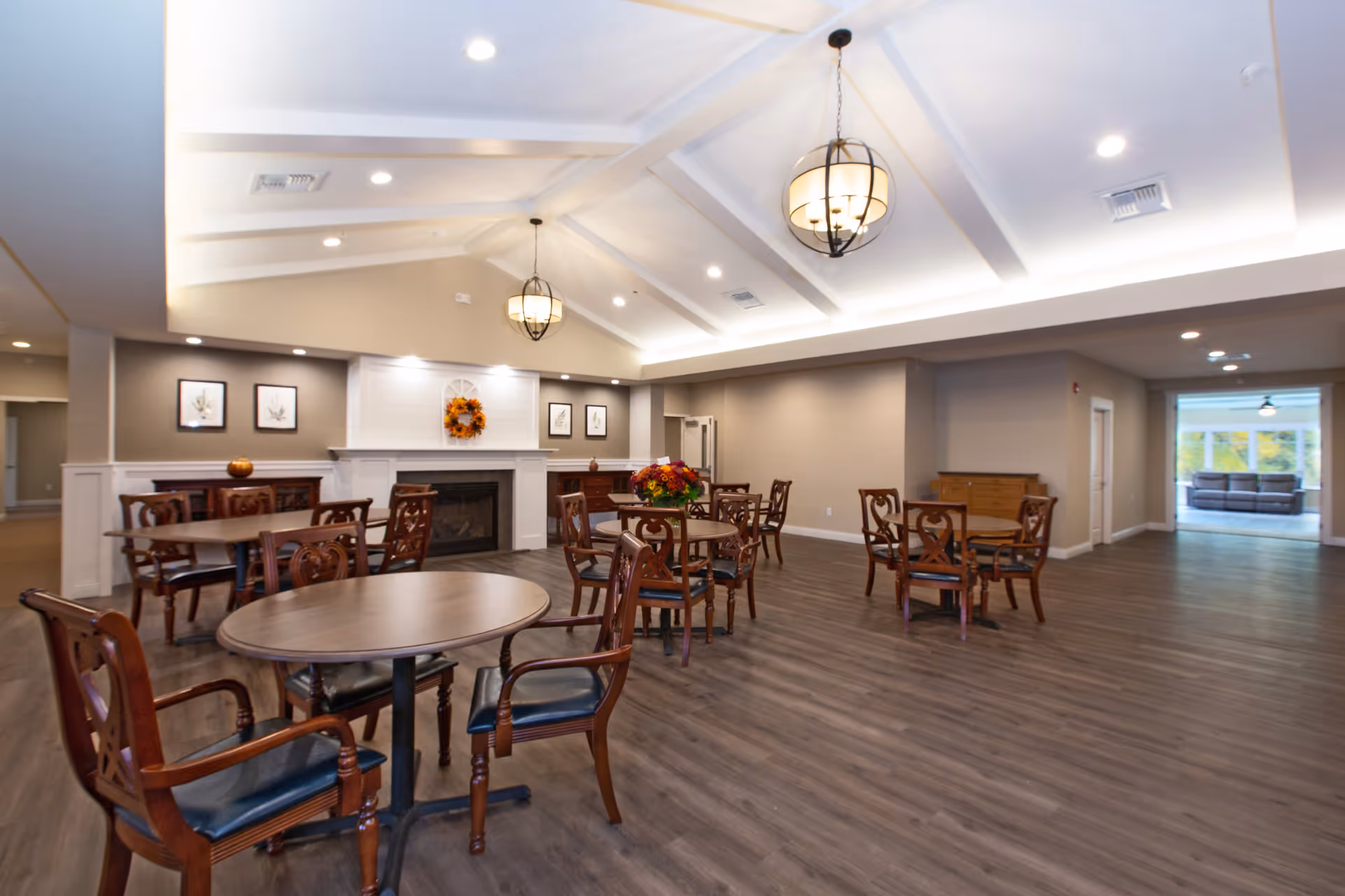 Spacious communal dining area with round wooden tables and chairs, a fireplace with a wreath, pendant lights, and a vaulted ceiling.