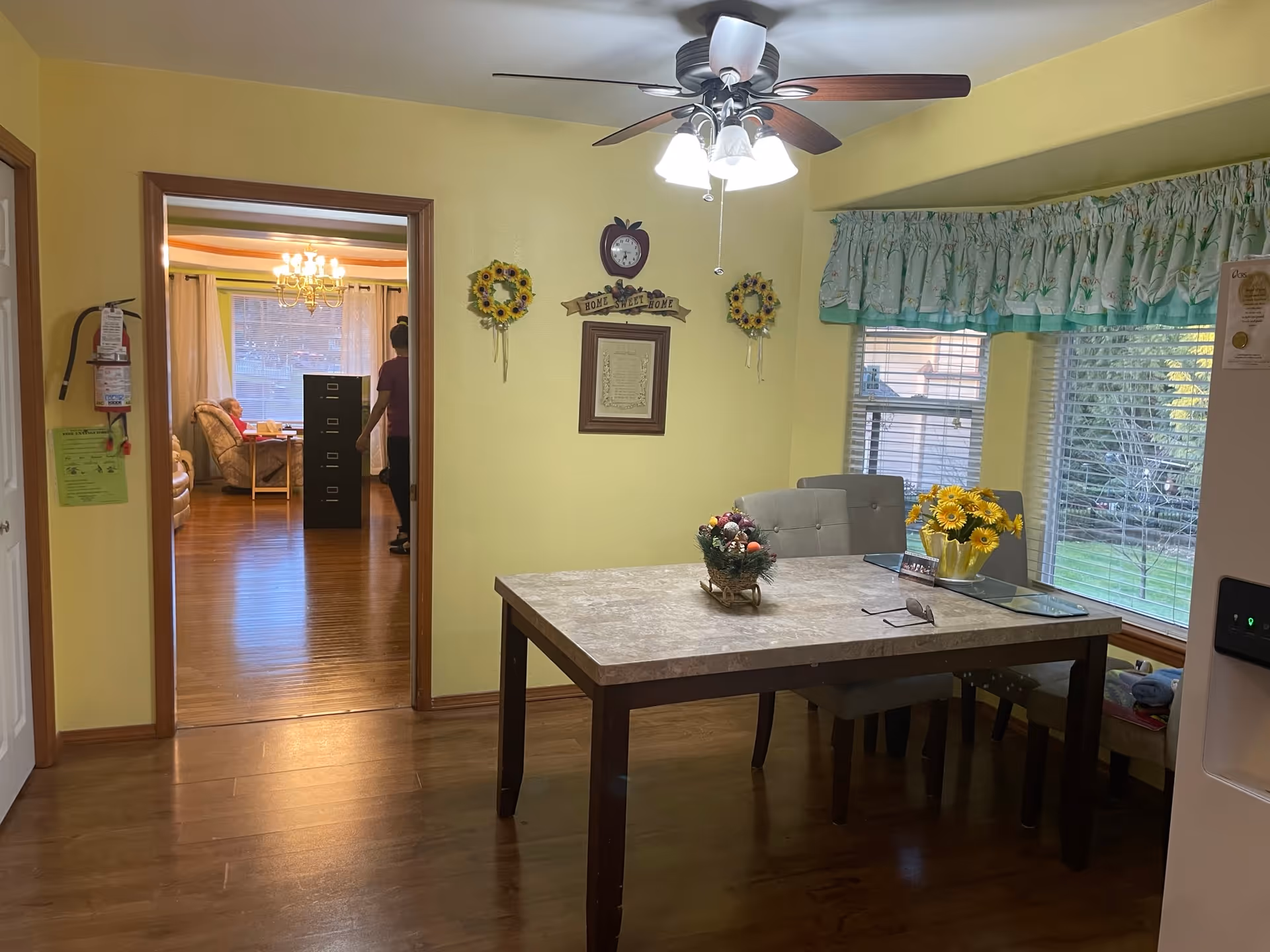 Dining room with a table and chairs, ceiling fan, window with floral valance, and a view into a living area through an open doorway.