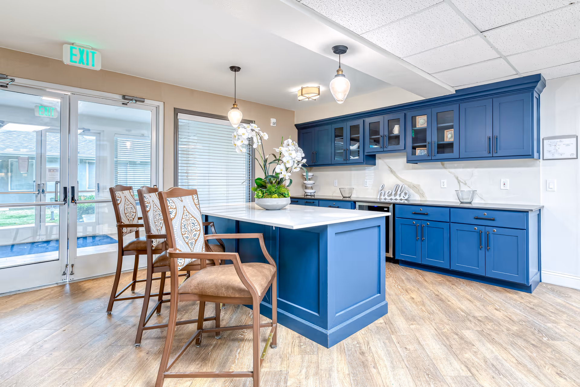 A bright kitchen area with blue cabinetry and a white countertop island. Three wooden chairs with patterned cushions are placed at the island. There are pendant lights hanging above the island, a decorative plant on the countertop, and glass doors leading outside. The floor is wood, and the walls are light-colored.