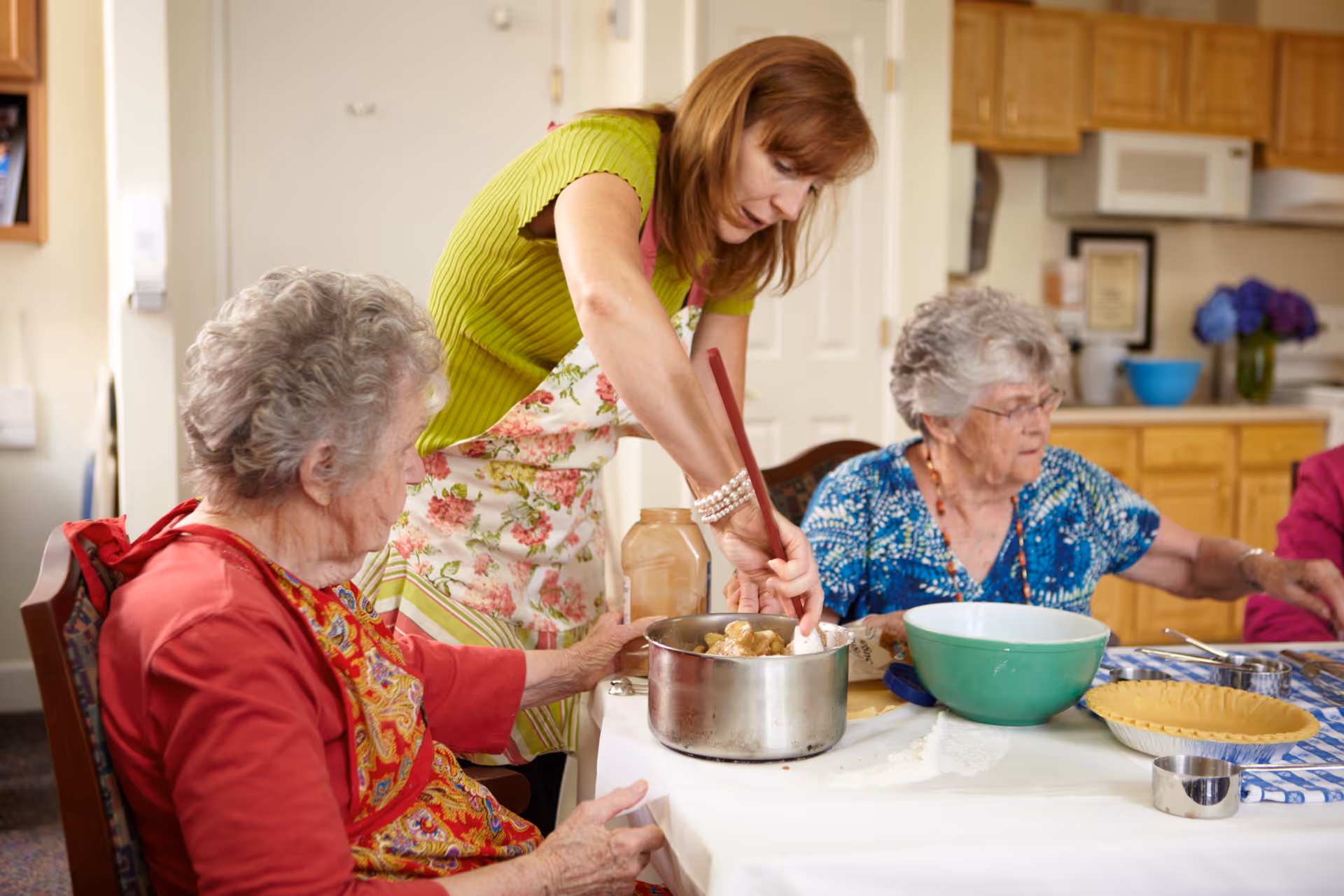 A woman in a green shirt and floral apron is stirring food in a pot on a table while two elderly women sit nearby, one in a red cardigan and the other in a blue patterned blouse, in a kitchen setting with wooden cabinets and a microwave in the background.