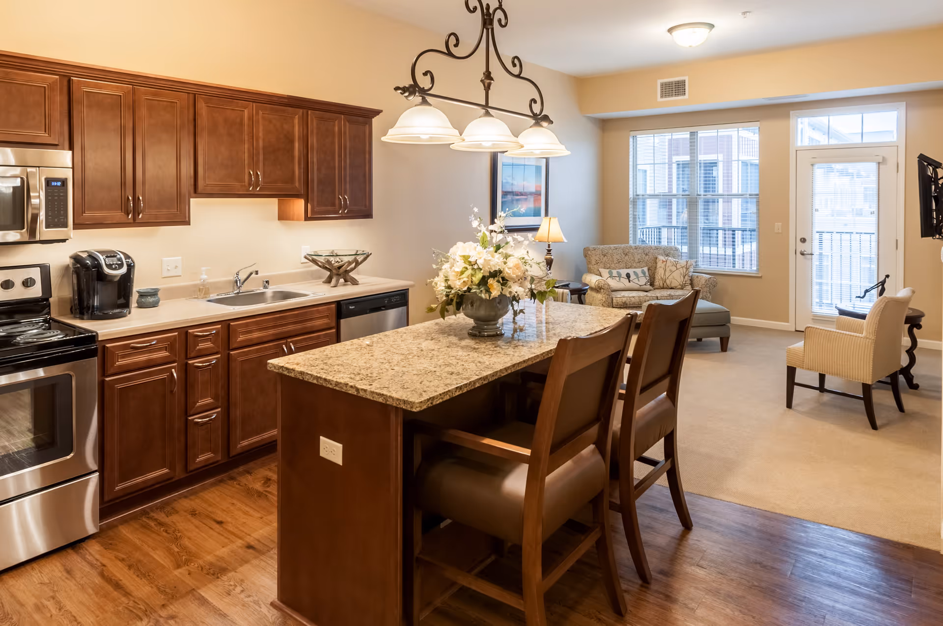 A bright and spacious kitchen and living area in a senior living facility. The kitchen features wooden cabinets, stainless steel appliances including a stove and microwave, a coffee maker, and a granite island with two chairs. A decorative light fixture hangs above the island. The living area has a patterned loveseat, an armchair, a side table with a lamp, a wall-mounted TV, and large windows with blinds letting in natural light.