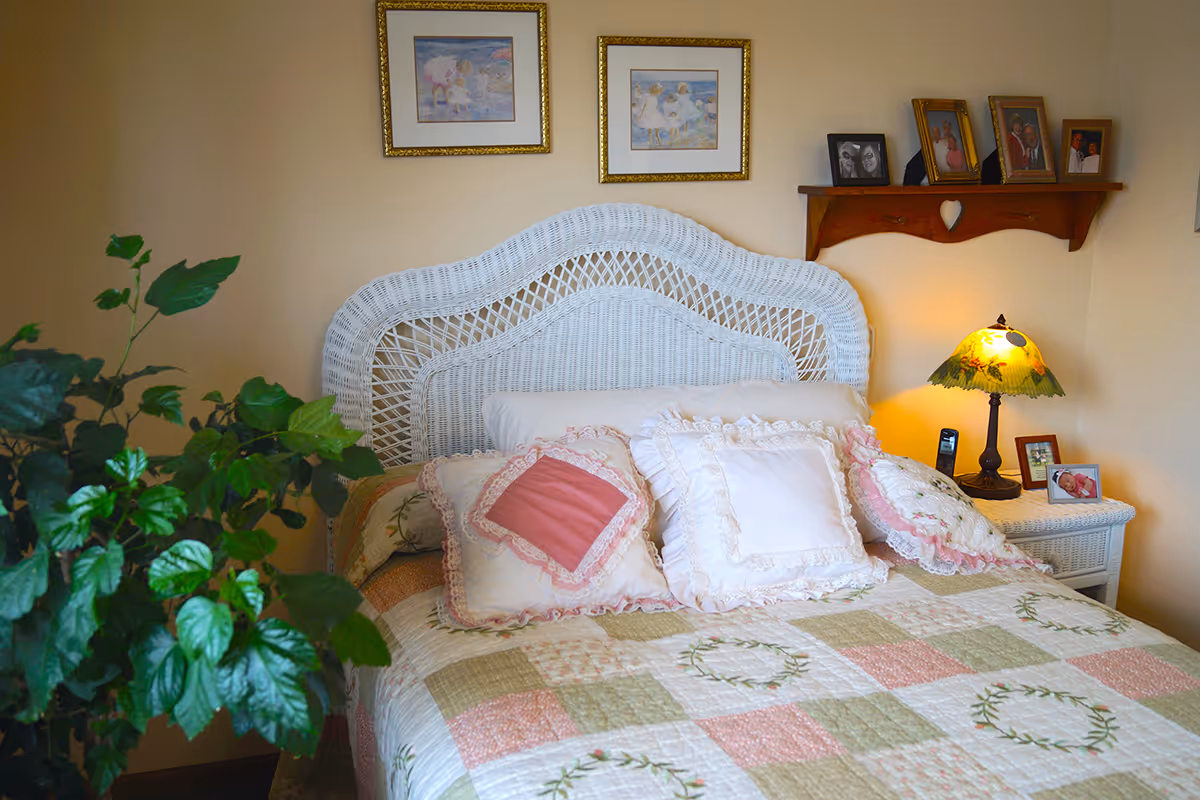 A cozy bedroom with a white wicker headboard bed adorned with multiple decorative pillows and a patchwork quilt in soft pastel colors. A green leafy plant is visible on the left side. On the right, there is a white wicker nightstand with a decorative lamp and framed photos. Above the nightstand, a wooden shelf holds more framed pictures. Two framed paintings hang on the beige wall above the bed.
