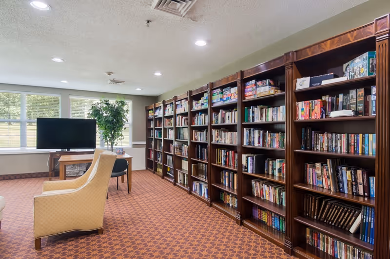 A cozy library room with multiple dark wooden bookshelves filled with books and board games along one wall. There are two beige upholstered chairs facing a flat-screen TV on a wooden stand near large windows letting in natural light. A green potted plant is placed near the TV, and the room has a patterned carpet and recessed ceiling lights.