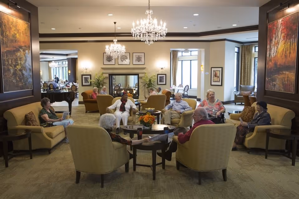 A group of elderly people sitting and conversing in a spacious, well-lit living room area with comfortable armchairs and sofas. The room features chandeliers, large windows, framed artwork on the walls, and a fireplace with decorative plants on either side.