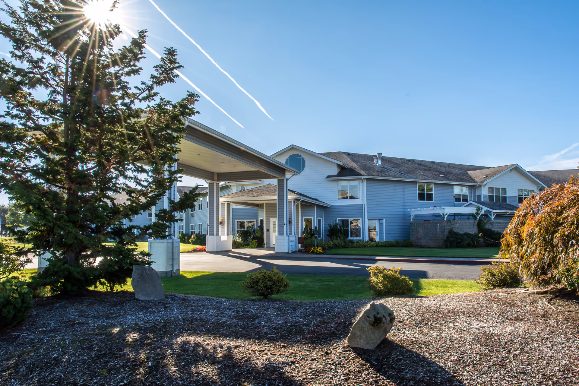 Front exterior of a two-story assisted living building with a porte-cochère entrance, landscaped grounds, and a bright sunny sky.