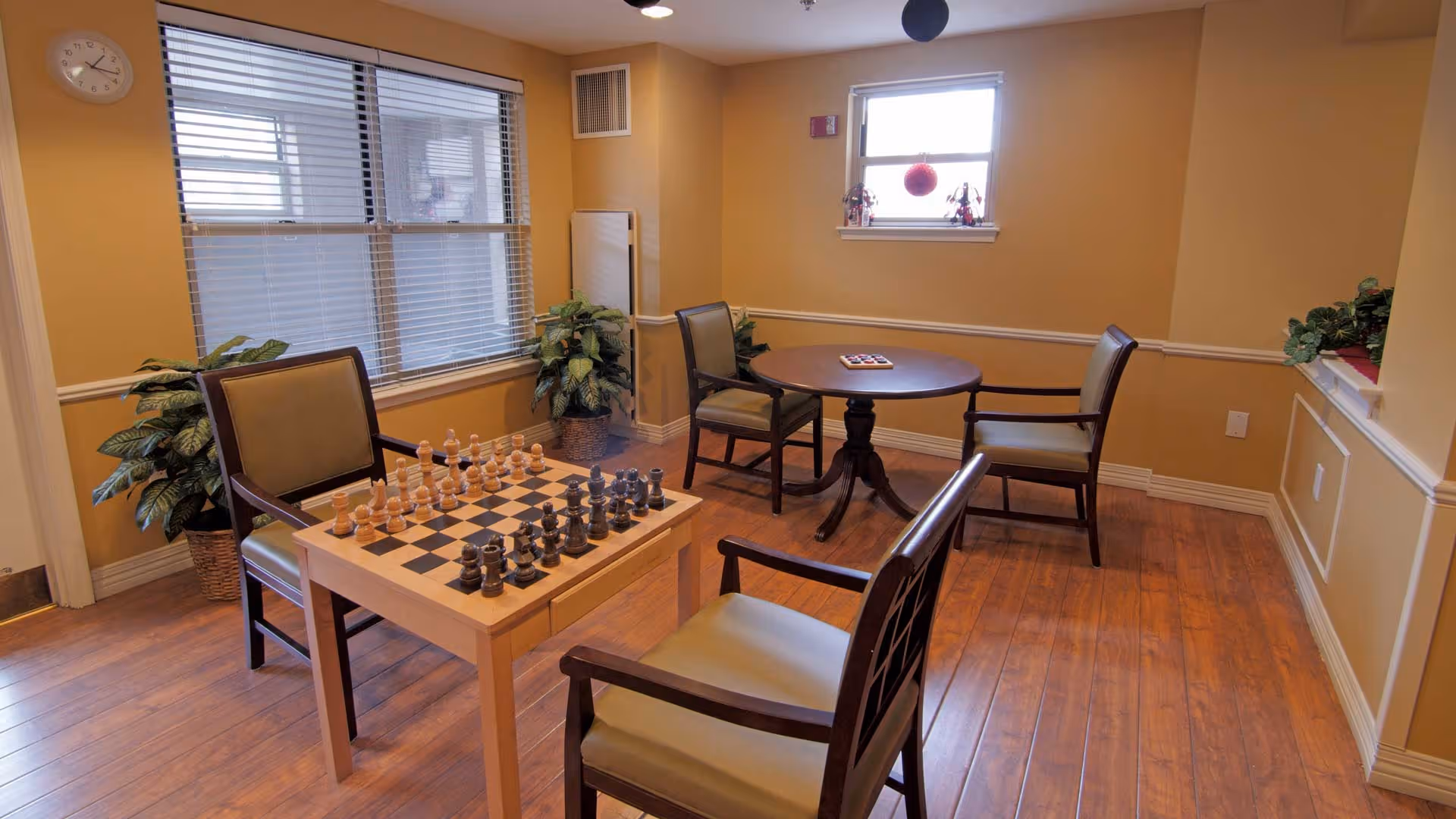 A cozy room with wooden flooring and beige walls featuring two seating areas. One area has a square table with a chessboard and chess pieces set up, surrounded by three chairs. The other area has a round table with two chairs. There are two windows, one large with blinds and one smaller with decorative items on the sill. Potted plants are placed near the walls, adding greenery to the space.
