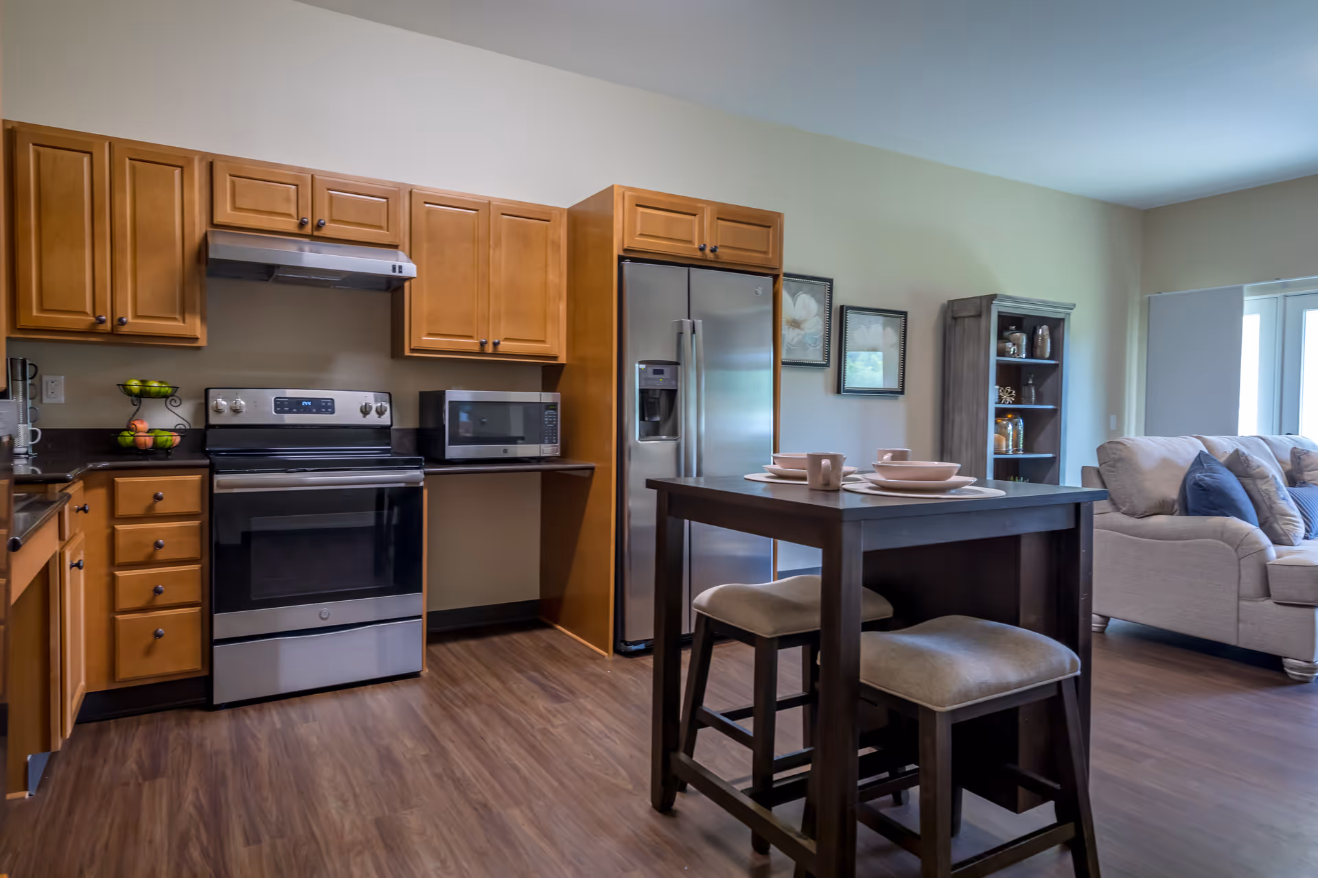 A modern kitchen and living area featuring wooden cabinets, a stainless steel stove and refrigerator, a microwave, and a small dining table with two cushioned stools. The living area includes a beige sofa with blue pillows and a wooden shelving unit with decorative items. The room has wood flooring and light-colored walls with framed artwork.