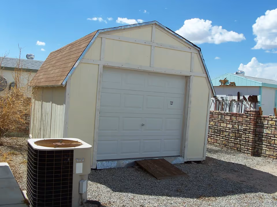 A small beige shed with a brown shingled roof and a white garage door, situated on a gravel surface. In front of the shed is an air conditioning unit. There is a brick wall and some other buildings in the background under a blue sky with scattered clouds.