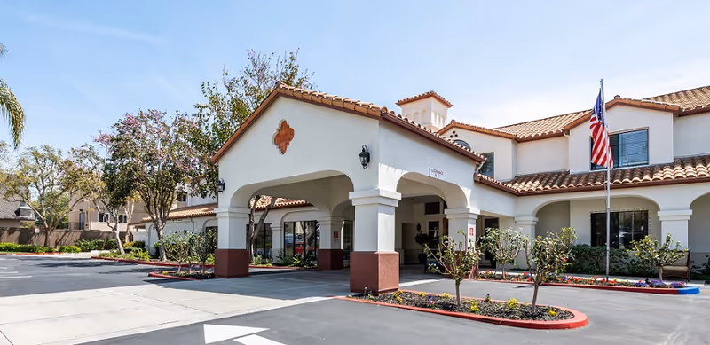 Exterior view of a senior living facility with a covered entrance, white walls, and a red-tiled roof. There are small landscaped areas with flowers and shrubs, an American flag on a flagpole, and a clear blue sky above.