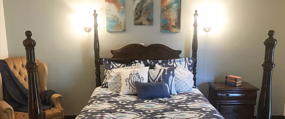 A bedroom featuring a wooden four-poster bed with patterned bedding and multiple pillows. On the left side, there is a tan upholstered armchair with a dark throw draped over it. On the right side, there is a wooden nightstand with a few books stacked on top. Two wall-mounted lights and two abstract paintings are visible above the bed.