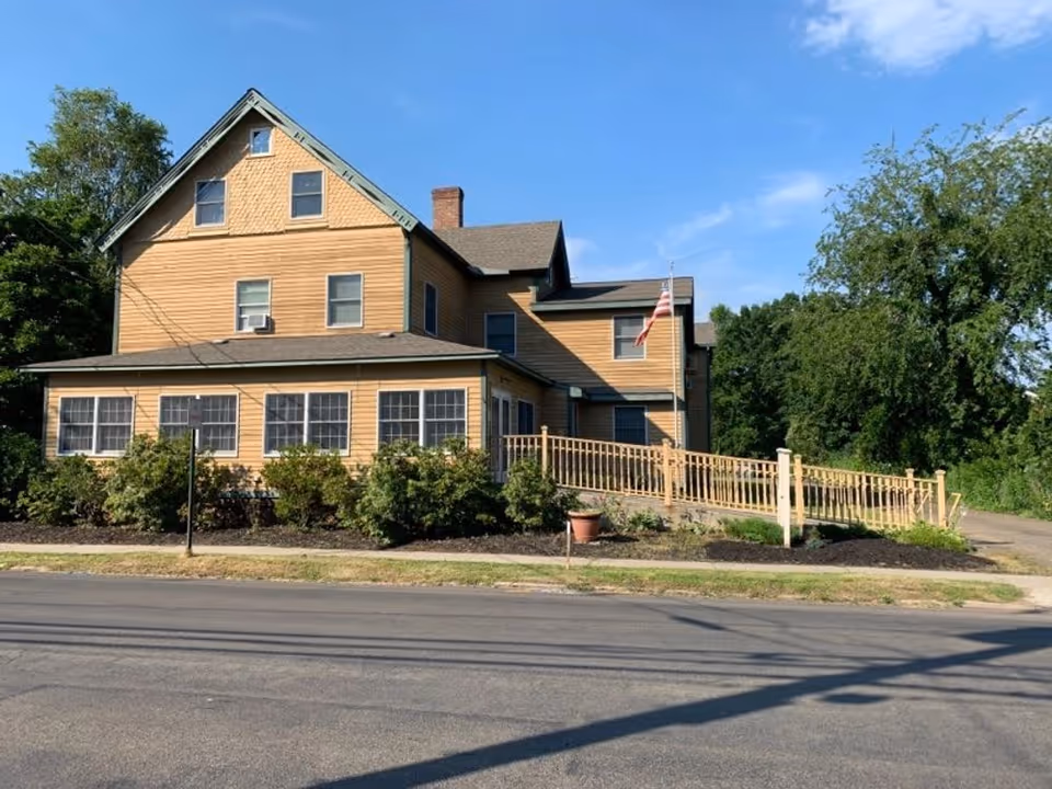 Exterior view of a large, multi-story beige house with multiple windows and a sloped roof. There is a wooden ramp leading to the entrance, surrounded by bushes and a small potted plant. An American flag is mounted near the entrance. The sky is clear and blue, and trees are visible in the background.