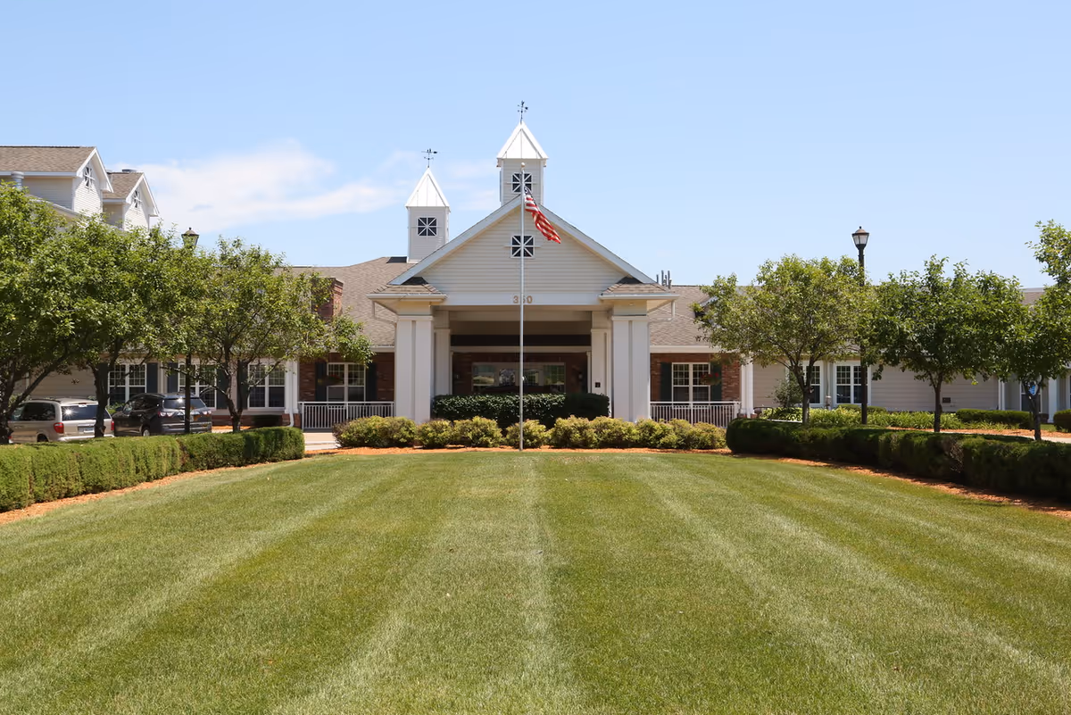 Front exterior view of Melrose Meadows Retirement Community building with a well-maintained lawn, trees lining the walkway, and an American flag on a flagpole in front of the entrance under a clear blue sky.